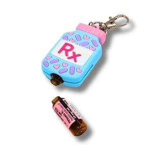 Peut inclure: Un porte-clés en silicone bleu clair en forme de flacon de médicament avec un bouchon rose, des cœurs roses et des pilules violettes. L'avant présente une étiquette blanche avec le texte Rx en rose. Une fiole en verre brun est en dessous.