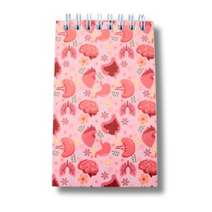 Peut inclure: Un bloc-notes à spirale avec une couverture rose ornée d'un motif répété d'organes humains stylisés, dont des cerveaux, des cœurs, des poumons, des reins et des estomacs, entrecoupés de petites fleurs. Le bloc-notes a un fond blanc.