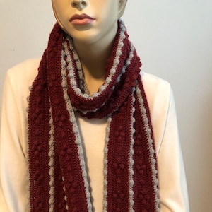 May include: A burgundy and gray knitted scarf. The scarf features a textured pattern with bobbles and a gray border. The scarf is draped around a mannequin's neck, showcasing its design and texture. The scarf is a winter accessory.
