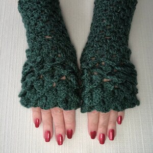 May include: A pair of dark green crochet fingerless gloves. The gloves have a textured, scale-like pattern and a ruffled edge at the wrist. The hands are visible, with red nail polish.