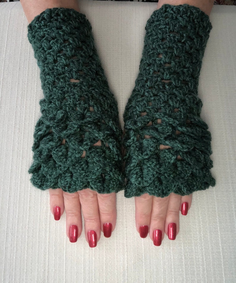 May include: A pair of dark green crochet fingerless gloves. The gloves have a textured, scale-like pattern and a ruffled edge at the wrist. The hands are visible, with red nail polish.