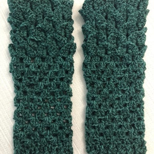 May include: Two teal green crochet fingerless gloves. The gloves have a textured, open weave pattern and a scalloped edge. The gloves are displayed on a white surface.