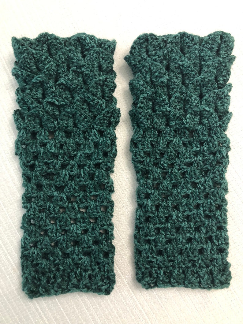 May include: Two teal green crochet fingerless gloves. The gloves have a textured, open weave pattern and a scalloped edge. The gloves are displayed on a white surface.