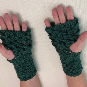 May include: A pair of dark green, fingerless gloves. The gloves are crocheted with a scale-like pattern, covering the back of the hands and wrists. The design features a hole for the thumb. The gloves are displayed against a neutral background.