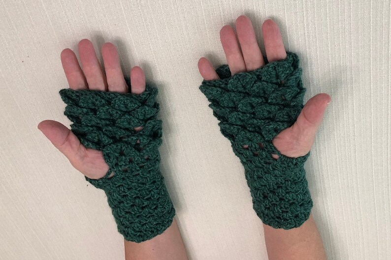 May include: A pair of dark green, fingerless gloves. The gloves are crocheted with a scale-like pattern, covering the back of the hands and wrists. The design features a hole for the thumb. The gloves are displayed against a neutral background.