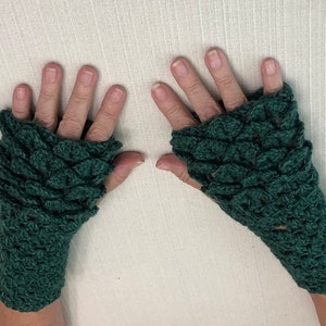 May include: A pair of dark green, fingerless crochet gloves. The gloves have a scale-like pattern covering the top of the hand and wrist. The gloves are made of a soft, textured yarn and are displayed on a light-colored surface.