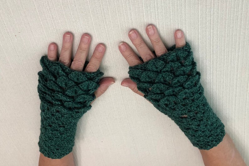 May include: A pair of dark green, fingerless crochet gloves. The gloves have a scale-like pattern covering the top of the hand and wrist. The gloves are made of a soft, textured yarn and are displayed on a light-colored surface.