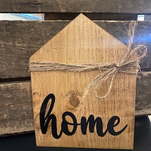 Könnte beinhalten: Ein hölzernes Schild in Hausform mit dem Wort "home" in schwarzer, kursiv geschriebener Schrift. Das Schild ist mit Schnur gebunden und hat einen rustikalen, ländlichen Stil.