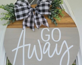 May include: A round wooden door hanger with a gray and wood background. The top half of the hanger is wood and the bottom half is gray. The words "GO AWAY (PLEASE)" are written in white on the gray background. A black and white checkered bow is attached to the top of the hanger with green leaves.