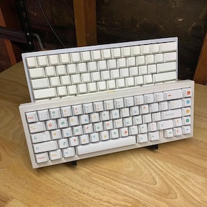May include: Two white mechanical keyboards with different keycaps. The top keyboard has blank white keycaps, while the bottom keyboard has colourful keycaps with letters and symbols. Both keyboards are on a wooden surface.