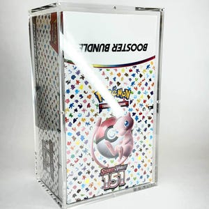 May include: A clear acrylic display case holding a Pokemon Trading Card Game Booster Bundle. The bundle features a colorful design with the text "BOOSTER BUNDLE" and "SCARLET & VIOLET 151" along with a pink Mew character.