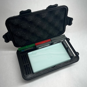 Könnte beinhalten: Geöffnete, schwarze Hartschalenkoffer mit einer hellblauen Nintendo DS Konsole und mehreren Spielkassetten. Der Koffer hat eine Schaumstoffauskleidung und sichere Verschlüsse. Entwickelt zum Schutz und zur Aufbewahrung von Gaming-Zubehör.