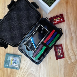 Puede incluir: Caja negra abierta que contiene una consola de juegos portátil plateada y lápices de colores. Se ven varios cartuchos de juego rojos, azules y verdes. Una jardinera blanca con musgo y un elemento decorativo se encuentra en el fondo.