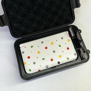 Könnte beinhalten: Eine weiße Nintendo 3DS Handheld-Spielekonsole mit einem benutzerdefinierten Animal Crossing-Design. Die Konsole befindet sich in einem schwarzen Hartschalenkoffer mit einem weichen schwarzen Innenraum. Der Koffer hat einen Verschluss an der Vorderseite und einen Griff oben.