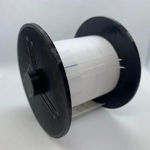 May include: A black plastic spool with a white label. The spool is used for holding and dispensing tape.