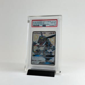 May include: A graded Pok&eacute;mon card featuring Rayquaza GX, from the 2018 Sun & Moon set. The card is encased in a clear protective holder and displayed on a black stand. The card's details include the set number, a grade of 10, and the text "Tempest GX".