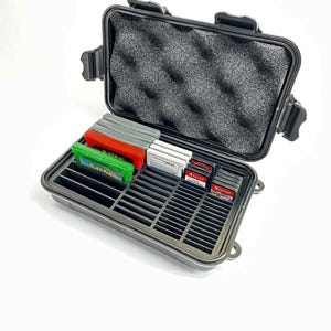 May include: A black, hard-shell case with a foam-lined top and a tray with slots for game cartridges. The case is open, revealing various game cartridges in red, green, and grey colours. The case is designed to protect and organise video game cartridges.