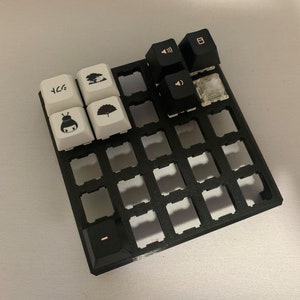 May include: A black plastic keyboard keycap holder with a grid of 36 slots. The holder has four white keycaps with Japanese characters and symbols, and three black keycaps with symbols.