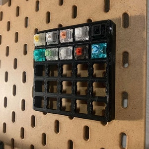 Puede incluir: Un organizador de plástico negro con una cuadrícula de ranuras para guardar teclas de teclado mecánico. El organizador tiene una variedad de teclas de diferentes colores, incluyendo verde, azul, blanco y naranja.