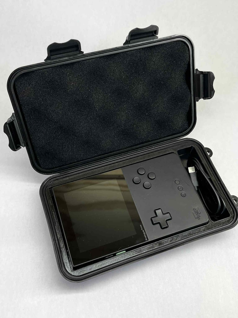 K&ouml;nnte beinhalten: Schwarze Handheld-Spielkonsole in einem offenen, schwarzen Schutzgeh&auml;use. Die Konsole verf&uuml;gt &uuml;ber ein Steuerkreuz, Tasten und einen Bildschirm. Ein USB-Kabel ist sichtbar. Das Geh&auml;use hat eine Schaumstoffauskleidung.