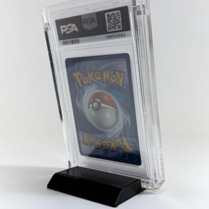 May include: A graded Pok&eacute;mon trading card encased in a clear plastic holder, displayed on a black stand. The card's reverse side is visible, featuring a swirling design and the Pok&eacute;mon logo. The holder has the PSA and PEA logos.