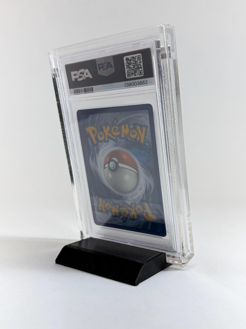 May include: A graded Pok&eacute;mon trading card encased in a clear plastic holder, displayed on a black stand. The card's reverse side is visible, featuring a swirling design and the Pok&eacute;mon logo. The holder has the PSA and PEA logos.