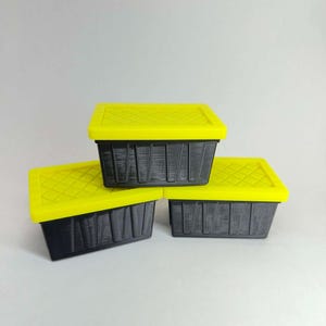 May include: Three black rectangular storage containers with bright yellow lids. The lids have a diamond pattern. The containers are stacked on a white surface. The containers are made of a textured material.