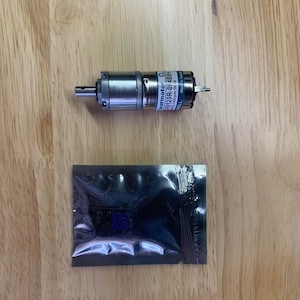 May include: A silver gearmotor with a black and blue label and a small, clear plastic package containing a blue component are displayed on a wooden surface. The gearmotor has a cylindrical shape with a protruding shaft.