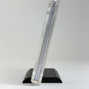 May include: A clear, rectangular photo frame with vertical ribbed detailing, resting on a black base. The frame is angled, showcasing its transparent design. The overall aesthetic is modern and minimalist, suitable for displaying photographs.