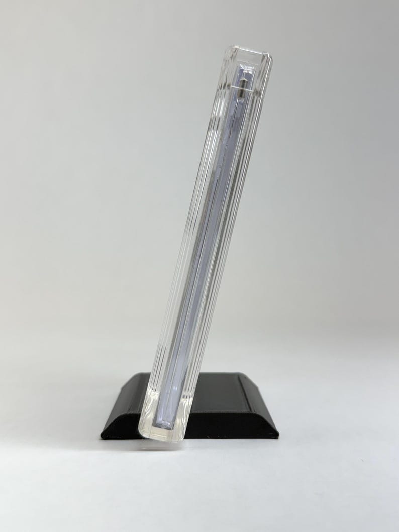 May include: A clear, rectangular photo frame with vertical ribbed detailing, resting on a black base. The frame is angled, showcasing its transparent design. The overall aesthetic is modern and minimalist, suitable for displaying photographs.