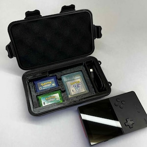 K&ouml;nnte beinhalten: Schwarzer Schutzkoffer mit drei Pok&eacute;mon Game Boy-Cartridges und einem schwarzen Handheld-Gaming-Ger&auml;t. Der Koffer ist ge&ouml;ffnet und zeigt blaue, gr&uuml;ne und transparente Cartridges. Das Handheld-Ger&auml;t ist schwarz mit rotem Akzent.