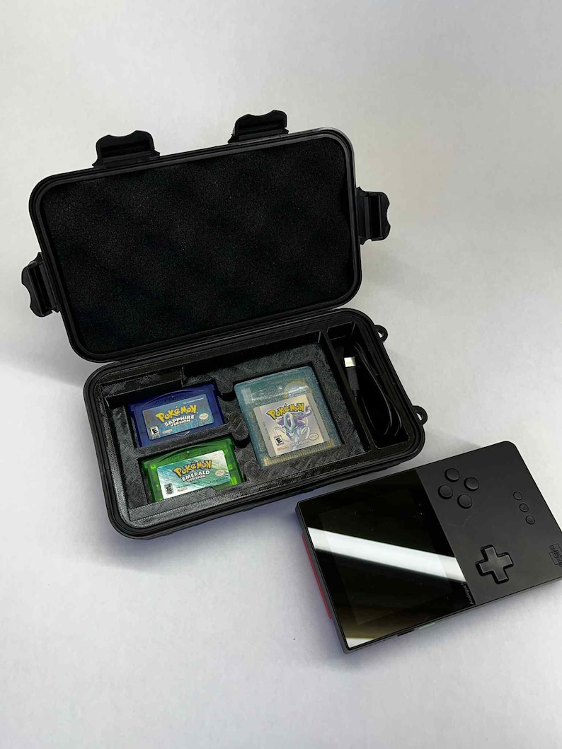 K&ouml;nnte beinhalten: Schwarzer Schutzkoffer mit drei Pok&eacute;mon Game Boy-Cartridges und einem schwarzen Handheld-Gaming-Ger&auml;t. Der Koffer ist ge&ouml;ffnet und zeigt blaue, gr&uuml;ne und transparente Cartridges. Das Handheld-Ger&auml;t ist schwarz mit rotem Akzent.