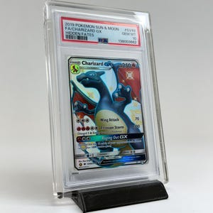 May include: A graded Charizard GX trading card from the 2019 Pokemon Sun & Moon Hidden Fates set. The card is encased in a clear plastic holder and displayed on a black stand. The card features a blue Charizard with red and orange accents.