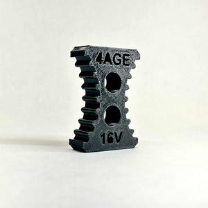 Op de afbeelding: Een zwart 3D-geprint object met de tekst "4AGE" en "16V". Het object heeft een unieke vorm met twee ronde gaten en een geribbeld ontwerp aan de zijkanten. Het staat tegen een effen witte achtergrond.