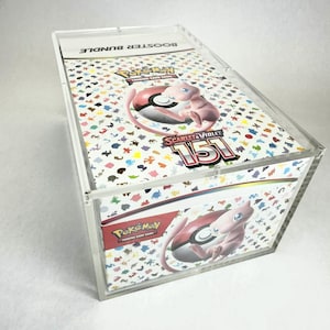May include: A clear acrylic box containing a Pokemon Trading Card Game booster bundle. The box features the "Scarlet & Violet 151" set with a pink Mew character. The top of the box reads "BOOSTER BUNDLE".