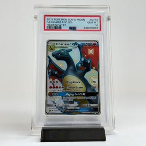 May include: A Charizard GX Pok&eacute;mon card from the Hidden Fates set, graded and encased in a protective holder. The card features a black and orange Charizard illustration with the text "Wing Attack" and "Crimson Storm". The card is displayed on a black stand.