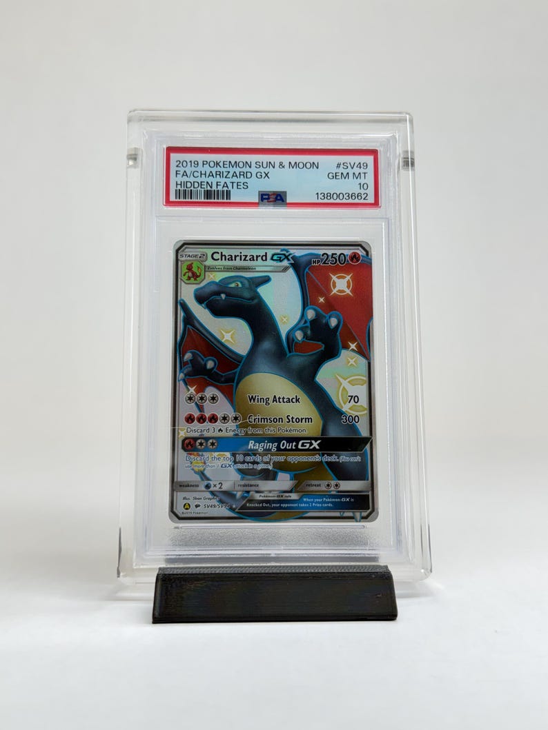May include: A Charizard GX Pok&eacute;mon card from the Hidden Fates set, graded and encased in a protective holder. The card features a black and orange Charizard illustration with the text "Wing Attack" and "Crimson Storm". The card is displayed on a black stand.