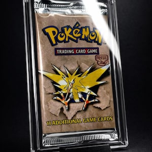 Può includere: Un pacchetto sigillato di carte collezionabili Pokémon del set Fossil, in una custodia protettiva trasparente. Il pacchetto presenta il logo Pokémon e un design di carta strappata che rivela una creatura gialla e nera. Il testo recita "11 ADDITIONAL GAME CARDS."