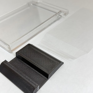May include: A clear plastic display case with a hinged lid, a black textured stand, and several clear plastic sleeves. The stand has a stepped design. The items are on a white surface.