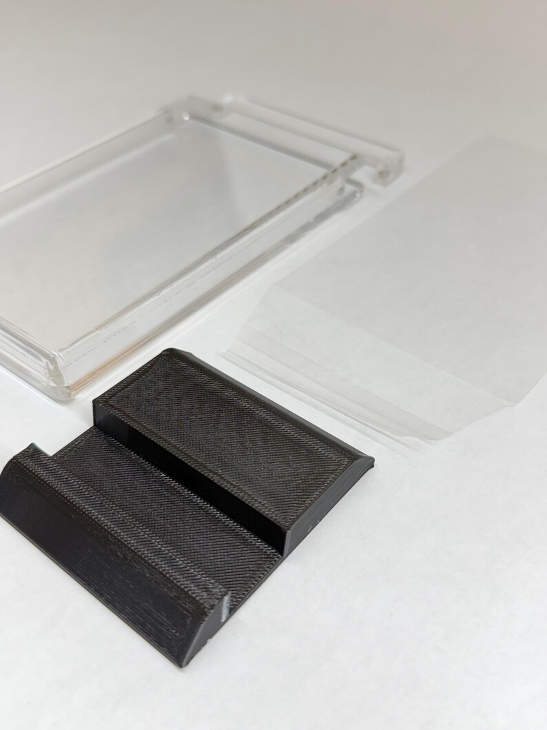 May include: A clear plastic display case with a hinged lid, a black textured stand, and several clear plastic sleeves. The stand has a stepped design. The items are on a white surface.
