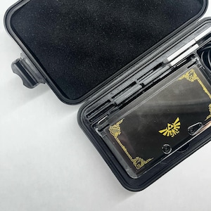 Può includere: Una custodia rigida nera con una console Nintendo 3DS XL dorata all'interno. La console ha un logo Triforce dorato sulla parte superiore.