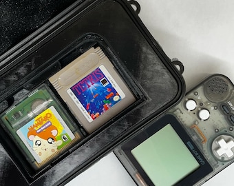 Op de afbeelding: Een zwarte harde koffer met twee Game Boy Color-cartridges, een met het spel "Hamtaro: Ham-Ham Games" en de andere met het spel "Tetris", en een transparante Game Boy Color-handheld.