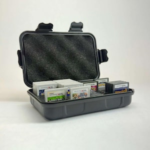 May include: A gray, rectangular case with a black interior, open to reveal multiple Nintendo 3DS game cartridges. The case has a foam insert in the lid and a secure latch. The games are labeled with various titles.