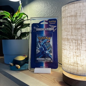 Può includere: Una teca di esposizione in plastica trasparente con all'interno un booster di gioco di carte collezionabili Pokemon Diamond & Pearl blu e bianco. Il booster presenta uno sfondo blu e bianco con un'immagine di due Pokemon, un Pokemon blu e bianco e un Pokemon bianco e blu. Il booster presenta anche il testo "Diamond & Pearl" e "10 carte di gioco aggiuntive".