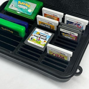 以下が含まれることがあります： さまざまなビデオゲームカートリッジが入った黒いプラスチック製のケース。ケースには側面にハンドルがあり、複数のカートリッジを収納できるように設計されています。カートリッジは、Nintendo 3DSとGame Boy Advanceのシステム用です。カートリッジには、ポケモンエメラルド、スーパーマリオブラザーズ、ポケモンパールなどのゲームが含まれています。