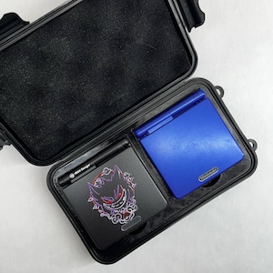 May include: A black hard case with a blue Game Boy Advance SP and a black Game Boy Advance SP with a purple and red graphic of a ghost-like creature. The case has a black interior and a black zipper.