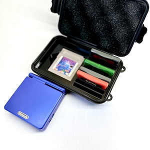 May include: A blue Nintendo Game Boy Advance SP console next to an open black case containing game cartridges. The case has a foam interior and is designed to protect the games. The cartridges are red, green, and gray.