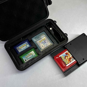 K&ouml;nnte beinhalten: Ein schwarzes Hartschalenetui mit einem Schaumstoffeinsatz enth&auml;lt drei Game Boy Advance-Cartridges: blaues Pok&eacute;mon Saphir, klares Pok&eacute;mon Silber und gr&uuml;nes Pok&eacute;mon Smaragd. Eine rote Pok&eacute;mon-Spielcartridge befindet sich in einem schwarzen Game Boy Advance SP.