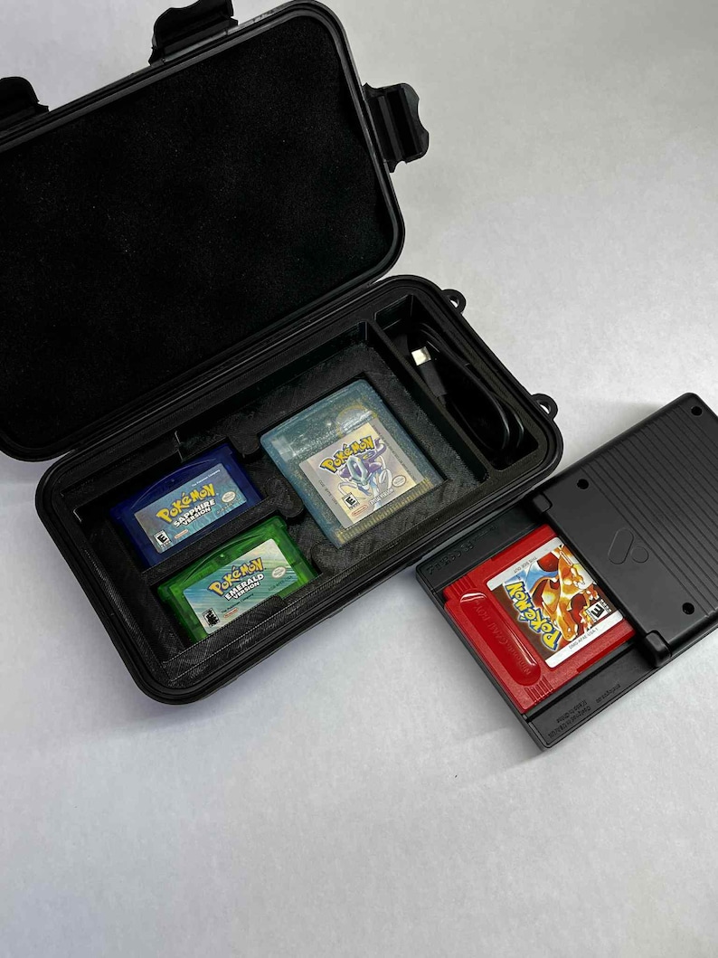 K&ouml;nnte beinhalten: Ein schwarzes Hartschalenetui mit einem Schaumstoffeinsatz enth&auml;lt drei Game Boy Advance-Cartridges: blaues Pok&eacute;mon Saphir, klares Pok&eacute;mon Silber und gr&uuml;nes Pok&eacute;mon Smaragd. Eine rote Pok&eacute;mon-Spielcartridge befindet sich in einem schwarzen Game Boy Advance SP.