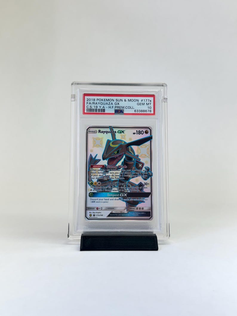 May include: A graded Rayquaza GX Pok&eacute;mon card from the 2018 Sun & Moon set. The card is encased in a clear protective holder and displayed on a black stand. The card features a green and blue Rayquaza illustration.